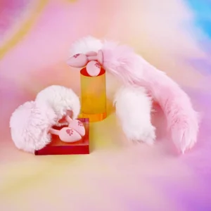 A playful arrangement of soft, fuzzy pink and white accessories, featuring a cute silicone anal plug with paw prints. The items are showcased against a vibrant, rainbow-colored background, with the anal butt plug tail resting on a red platform and surrounded by fluffy tails, creating a whimsical and inviting atmosphere.
