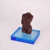 An elegant brown candle shaped like a stylized female torso, displayed on a striking blue acrylic base. The candle captures intricate details of the figure, with one arm gently cradling the body, creating an intimate and sophisticated aesthetic. The base contrasts beautifully with the rich color of the candle, enhancing its artistic presence. This piece is not just a candle, but a statement decor item, ideal for creating a romantic ambiance in any space, perfectly embodying the allure of sensual candles. The soft lighting casts gentle shadows, adding depth to the overall composition.