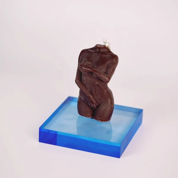 An elegant brown candle shaped like a stylized female torso, displayed on a striking blue acrylic base. The candle captures intricate details of the figure, with one arm gently cradling the body, creating an intimate and sophisticated aesthetic. The base contrasts beautifully with the rich color of the candle, enhancing its artistic presence. This piece is not just a candle, but a statement decor item, ideal for creating a romantic ambiance in any space, perfectly embodying the allure of sensual candles. The soft lighting casts gentle shadows, adding depth to the overall composition.