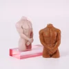 Two artistic candles shaped like headless male torsos, showcasing a range of skin tones. The lighter candle stands on a transparent pink acrylic base, capturing intricate details of well-defined muscles with one hand resting at its waist. Next to it, a darker-toned candle exhibits a similarly muscular form, arms crossed in front, adding a sense of elegance. Both candles are designed to provide a warm glow when lit, doubling as striking decor pieces. These unique creations embody the allure of sensual candles, perfect for enhancing any romantic atmosphere. The minimalist background emphasizes their artistic features.