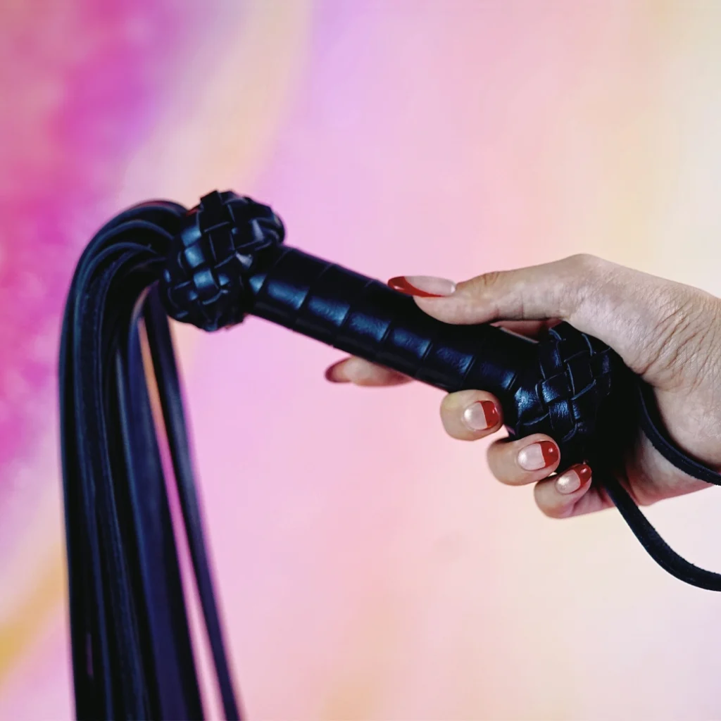 PB0305 A close-up of a beginner role play paddle from a whip kit is held in a hand against a soft, pastel gradient background. The paddle features a braided black handle with textured grips and decorative knots at each end, showcasing an elegant design. Long, sleek tails extend from the handle, flowing gracefully. This stylish beginner whip emphasizes comfort and accessibility, making it perfect for those exploring role play for the first time.