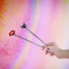 A hand holds two playful beginner role play paddle against a vibrant, gradient background in pastel colors. The paddles are crafted from stainless steel and feature decorative tops: one is a bold red lip shape, while the other is a cute black cat face with white whiskers. The sleek, shiny handles add an elegant touch, making these paddles both stylish and beginner-friendly for those exploring role play.