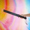 A hand holds a stylish beginner BDSM spanking paddle against a vibrant, colorful gradient background. The paddle features a textured black surface adorned with metallic studs along one edge for added flair. A smooth, ergonomic handle is accentuated with decorative detailing, providing a comfortable grip. The playful and elegant design of the paddle makes it an ideal choice for those new to BDSM play, inviting exploration and creativity.
