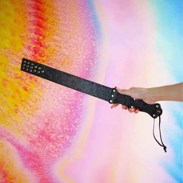 A hand holds a stylish beginner BDSM spanking paddle against a vibrant, colorful gradient background. The paddle features a textured black surface adorned with metallic studs along one edge for added flair. A smooth, ergonomic handle is accentuated with decorative detailing, providing a comfortable grip. The playful and elegant design of the paddle makes it an ideal choice for those new to BDSM play, inviting exploration and creativity.