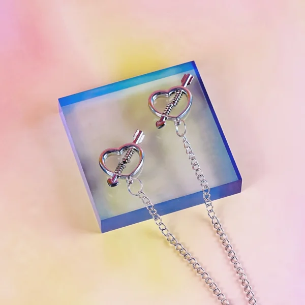 A pair of silver heart-shaped nipple clamps with chain, featuring an elegant arrow piercing through the heart design. The adjustable screw mechanism ensures customizable pressure, while the delicate connecting chain adds a sensual touch. Perfect for those exploring intimate accessories with a stylish and playful aesthetic.