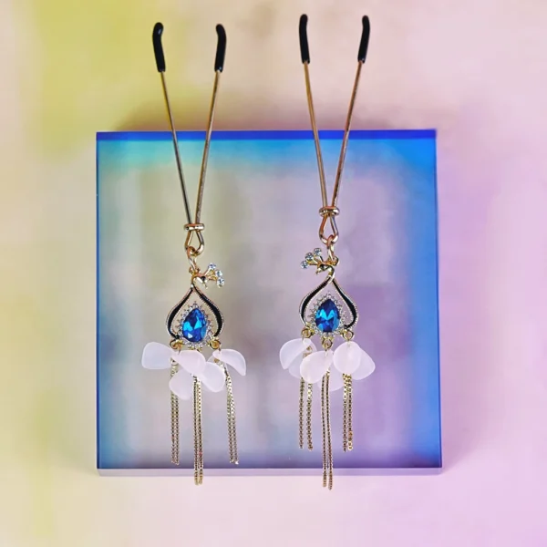 A pair of elegant BDSM nipple clamps is showcased against a softly illuminated, gradient background. The clamps feature delicate gold chains with intricate designs, adorned with vibrant blue gemstones and white floral accents. Rubber tips at the ends ensure comfort, while the dangling elements add a touch of glamour. This visually striking design makes them perfect for those exploring BDSM dynamics with a stylish flair.