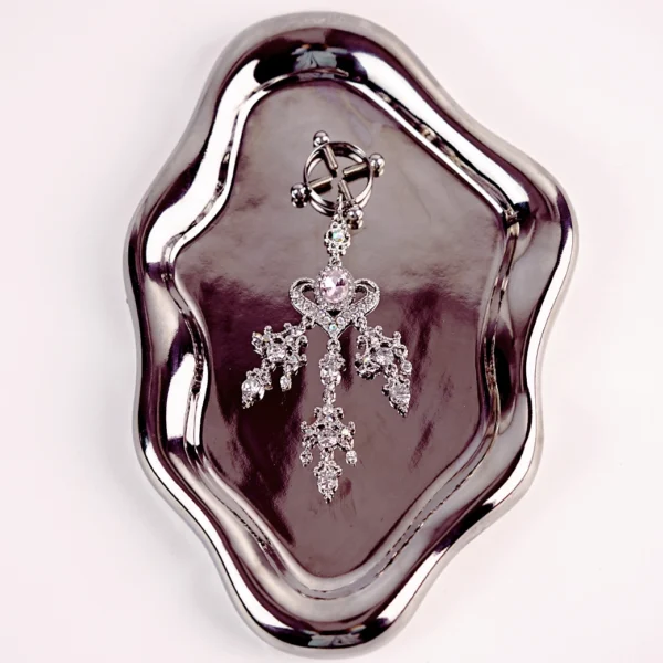 A luxurious clamp is elegantly displayed on a shiny, metallic tray. The clamp features an intricate design with silver detailing and sparkling gemstones, including a central lavender stone surrounded by shimmering accents. The clamp is styled with leaf-like embellishments that cascade down, adding a touch of glamour. This visually stunning piece combines functionality with elegance, perfect for those exploring role play dynamics.