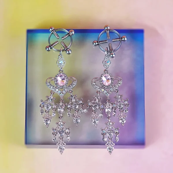 A pair of luxurious clamps is elegantly displayed against a gradient background. The clamps feature intricate silver designs, adorned with sparkling rhinestones, including a large central stone that adds a touch of glamour. Each clamp is finished with decorative dangling chains that create a cascading effect, enhancing their allure. This combination of beauty and function makes them perfect for those exploring role play dynamics with style.