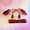 A vibrant display of burgundy bed restraints set against a soft, pastel background. The set includes two matching wrist cuffs with gold buckles, connected by a central strap that crosses elegantly in the middle. The cuffs feature decorative studs, enhancing their style and sophistication. Below, a flat strap lies ready for attachment, also adorned with gold hardware. This stylish collection showcases an inviting combination of functionality and aesthetics, making it a perfect choice for those exploring the playful side of bed restraints set. The light reflects beautifully off the leather, emphasizing the quality of the materials.