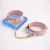 A playful pair of pink leather handcuffs designed for a stylish electro-stimulation bed restraints set. Each cuff is adorned with the words "PASSION" and "LOVE" in a subtle, textured finish, positioned around the outer edge. The cuffs feature gold metal hardware, including adjustable buckles and a connecting chain that includes a sturdy clip. Positioned against a bright white background, the cuffs rest on a vibrant blue acrylic stand, enhancing their eye-catching design. This charming accessory is a perfect example of bed restraints set, combining elegance and functionality for adventurous intimate experiences.