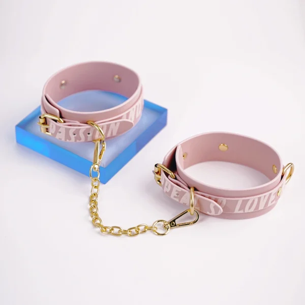 A playful pair of pink leather handcuffs designed for a stylish electro-stimulation bed restraints set. Each cuff is adorned with the words "PASSION" and "LOVE" in a subtle, textured finish, positioned around the outer edge. The cuffs feature gold metal hardware, including adjustable buckles and a connecting chain that includes a sturdy clip. Positioned against a bright white background, the cuffs rest on a vibrant blue acrylic stand, enhancing their eye-catching design. This charming accessory is a perfect example of bed restraints set, combining elegance and functionality for adventurous intimate experiences.