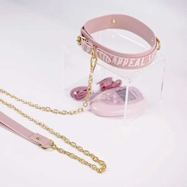 A stylish pink collar from an electro-stimulation bed restraints set, featuring the words "PASSION APPEAL" elegantly embossed along the strap. The collar is crafted from soft leather and is secured with gold metal buckles for a snug fit. Attached to the collar is a gold chain that connects to a matching leash, showcasing a combination of functionality and chic design. A pink control unit is partially visible in the background, hinting at the collar's electro-stimulation features. Displayed on a clear acrylic stand against a clean white background, this striking piece exemplifies modern bed restraints set, perfect for exploring intimate adventures with style.