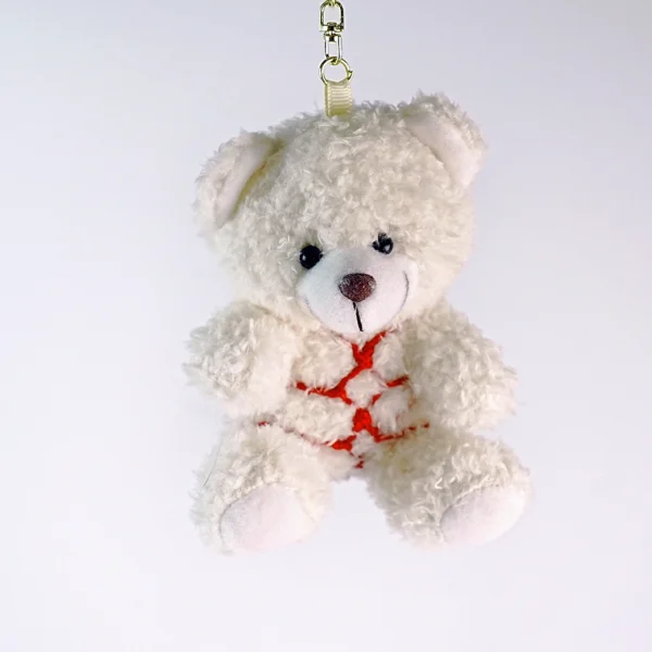 A cute white teddy bear designed as a bondage bear, featuring fluffy fur and a friendly face. The bear is adorned with a bright red harness that wraps around its body in a playful crisscross pattern. It is suspended with a clip at the top, allowing it to be easily attached to bags or keychains. Set against a clean white background, this charming and whimsical design perfectly represents the fun and lighthearted spirit of a bondage bear, making it an ideal accessory for collectors or those looking to add a playful touch to their items.