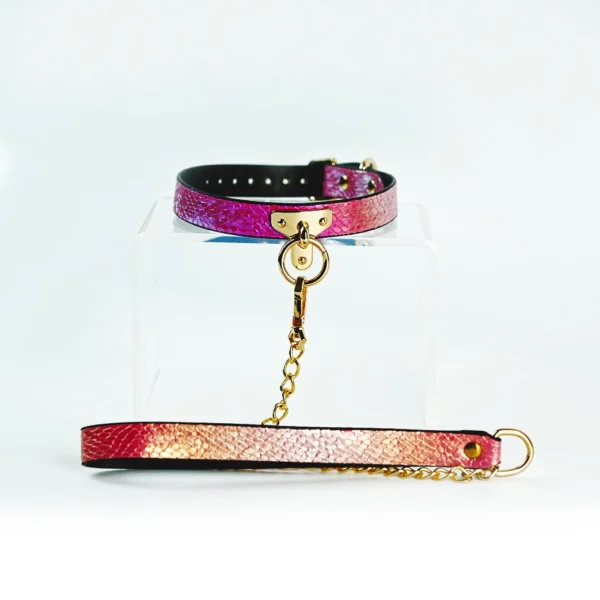 A vibrant restraint collar featuring a stunning gradient design that transitions from deep pink to shimmering gold. The collar is made of textured leather, elegantly showcasing a unique snakeskin pattern. It is secured with a gold metal buckle and includes a central D-ring, perfect for attaching a leash or other accessories. A matching leash, adorned with a similar gradient and a gold chain, extends from the collar, enhancing its stylish appearance. Displayed on a clear acrylic stand against a soft white background, this eye-catching piece represents modern restraint collar gear, ideal for those looking to explore intimacy with flair and sophistication.