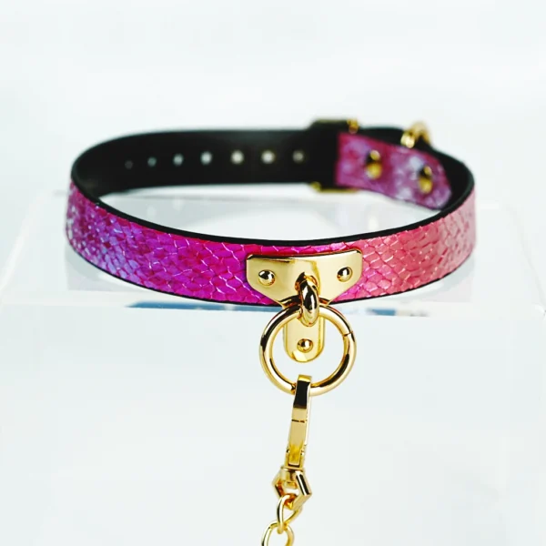 A vibrant restraint collar featuring a beautiful gradient of pink hues, transitioning to a shimmering gold. The collar is crafted from high-quality leather with a textured snakeskin design. It is secured with a gold metal buckle and showcases a central D-ring for versatility, perfect for attaching a leash or other accessories. A matching gold chain extends from the D-ring, adding an elegant touch. Displayed on a clear acrylic stand against a soft white background, this stunning piece exemplifies stylish restraint collar gear, ideal for those looking to explore intimacy with a blend of elegance and playfulness.