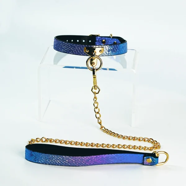 A stunning restraint collar displayed on a clear acrylic stand, featuring a vibrant blue and purple gradient with a textured snakeskin pattern. The collar is made of high-quality leather, secured with a gold metal buckle and a central D-ring. Attached to the D-ring is a sleek gold chain that connects to a matching leash, enhancing the overall aesthetic. This elegant piece of restraint collar gear combines sophistication with playful elements, perfect for those looking to explore intimate and adventurous experiences in style.