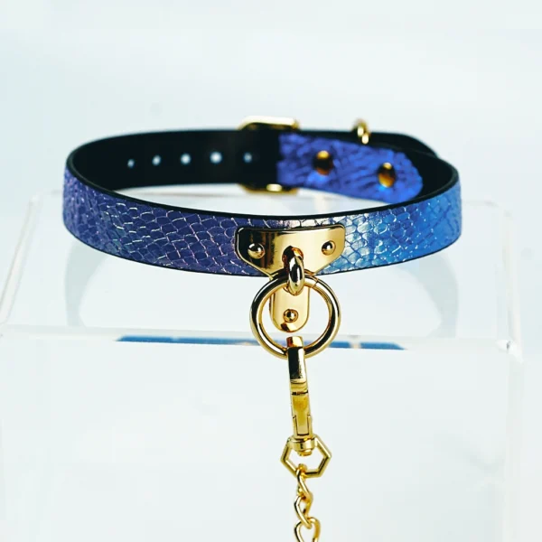 A stunning restraint collar featuring a vibrant blue and purple gradient with a textured snakeskin finish. The collar is crafted from high-quality leather and is secured with a sleek gold metal buckle. At the front, a central D-ring provides a stylish attachment point, leading to a decorative chain that adds an element of sophistication. The collar is displayed on a clear acrylic stand against a soft background, perfectly showcasing this elegant piece of restraint collar gear, ideal for those looking to explore intimate adventures with flair.