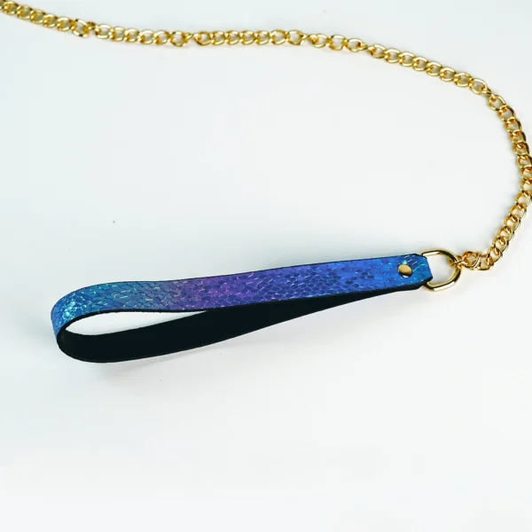 A close-up of a stylish BDSM collar attachment featuring a glossy blue and purple snakeskin-patterned strap that leads to a shiny gold chain. The strap has a black underside for added comfort, with a polished gold ring at the end for easy connection to a collar or leash. Displayed against a clean white background, this eye-catching accessory perfectly embodies modern restraint collar gear, combining playful elegance with functionality for those exploring intimate adventures.